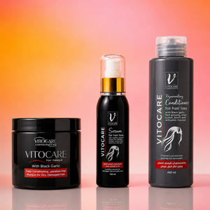 Three Vitocare hair care products arranged on a reflective surface against a warm orange-to-pink gradient background: a black jar labeled 'With Black Garlic,' a black pump bottle labeled 'Serum,' and a tall black bottle labeled 'Recovering Conditions for hair loss.'.