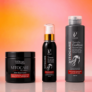 Three Vitocare hair care products arranged on a reflective surface against a warm orange-to-pink gradient background: a black jar labeled 'With Black Garlic,' a black pump bottle labeled 'Serum,' and a tall black bottle labeled 'Recovering Conditions for hair loss.'.
